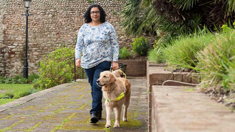 Lena and her guide dog, Alex, walk along a pathway toward the camera. Lena and her guide dog, Alex, walk along a pathway toward the camera.