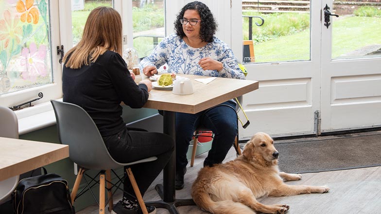 Lena sits in a café with her friend, as her guide dog sits at her feet.
