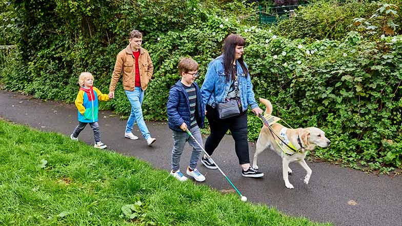 Lorna, her husband, and her two children, walk along a path. Lorna is guided by her guide dog.
