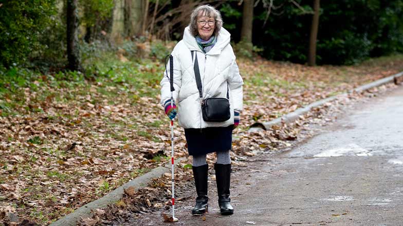 Hilary smiles as she stands on a wooded pathway, holding her long cane.