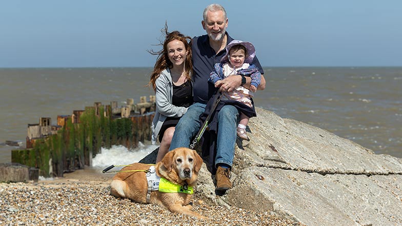 Simon, his daughter and granddaughter sitting closely together on a windy day with guide dog Mayne on a stony beach.