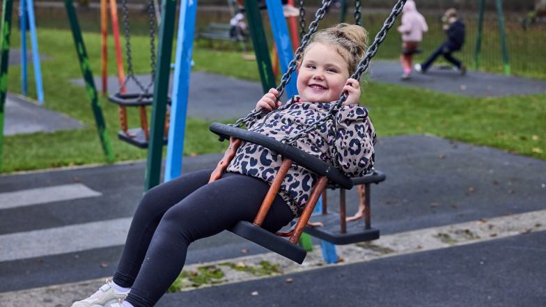 Mabel is smiling at the camera as she uses a swing in a playground