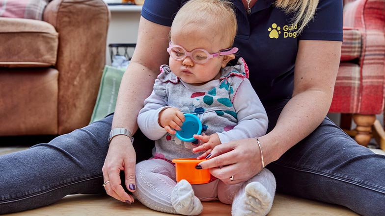 A young service user sits on the floor playing with toys, with her Habilitation Specialist behind her.