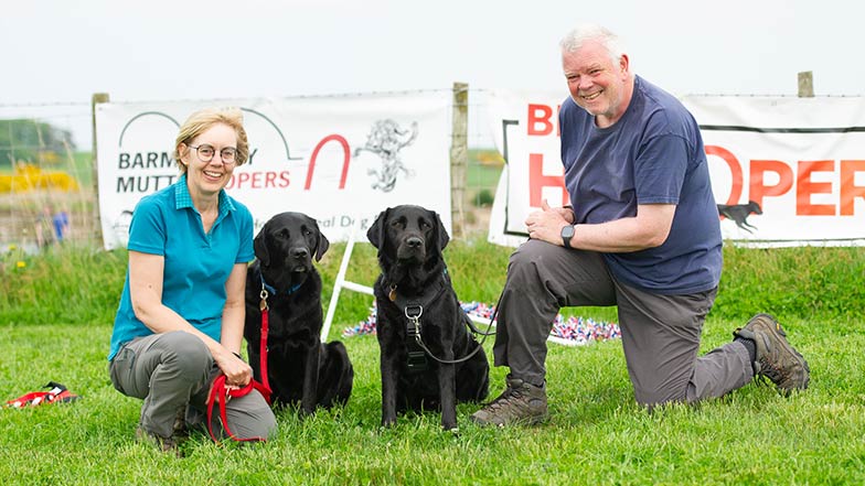 Cody, a rehomed dog, Rebus, Iain, and Jane, sit in front of signage for Canine Hoopers.