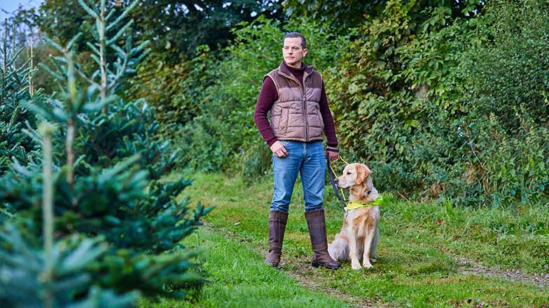 Scott and guide dog Milo standing together in a forest with a Christmas tree in the foreground