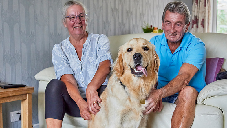 Volunteer fosterers Terry and Margaret sit on the sofa with guide dog Elwell before them poking his tongue out