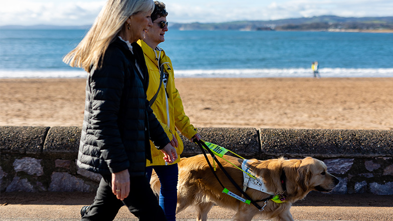 Volunteer Jan guides Suzanne and her guide dog across the sea front, the beach and sea are in the background