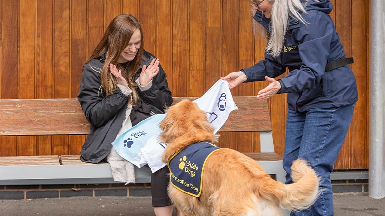 A demonstration dog performs a chin rest on a woman's lap as a trainer watches. They are all smiling.