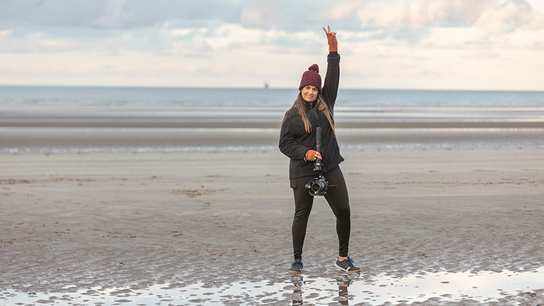 A Video Producer standing on a beach holding a camera and doing a peace pose