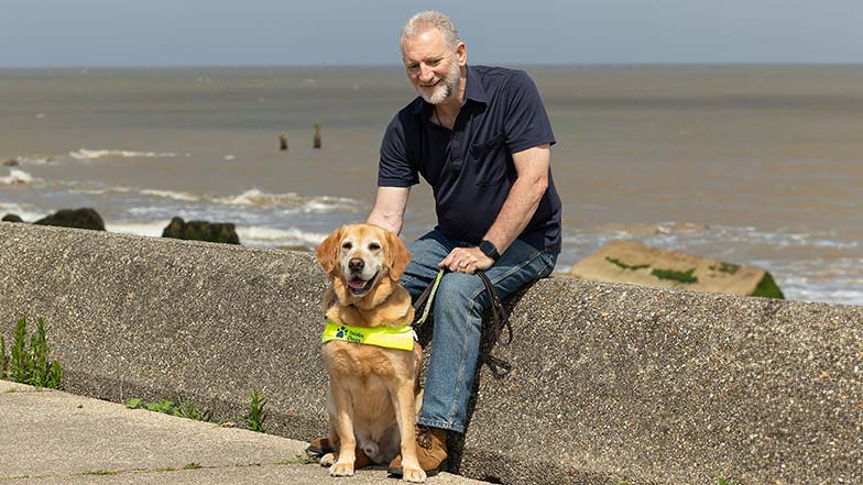 Guide dog owner Simon sitting on a sea wall with his guide dog sitting at his feet.