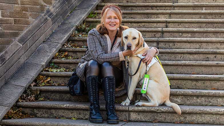 Heather sitting with guide dog Connor on a step at the bottom of an outdoor staircase. Heather has her arms around Connor and is smiling broadly.