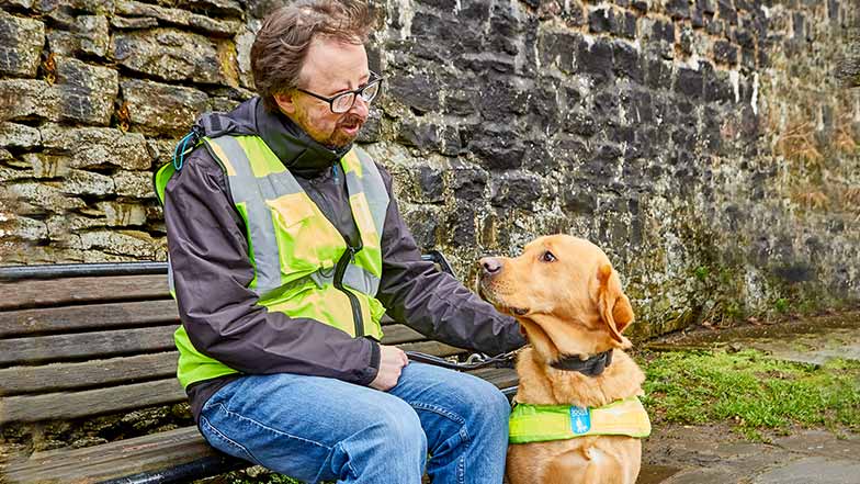 Guide dog owner Richard in a high vis vest sitting on a bench with his guide dog Martin at his feet