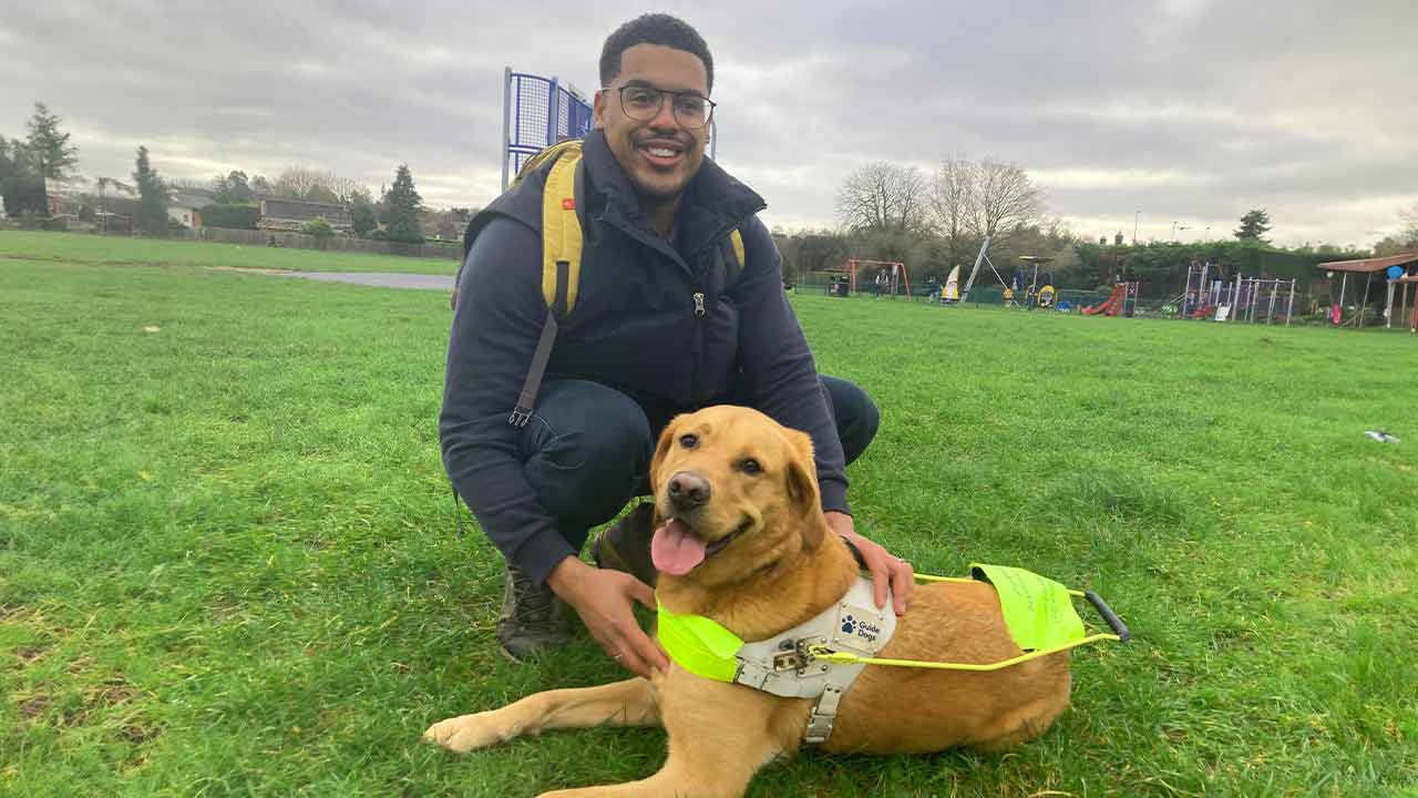 Guide dog owner, Devante, sits in a field with his guide dog.