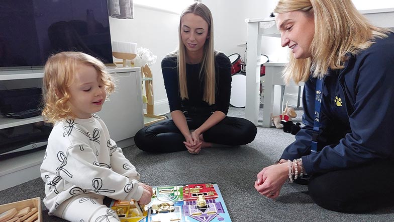 Eliza sits on the floor, smiling, with her mum and Habilitation Specialist. 