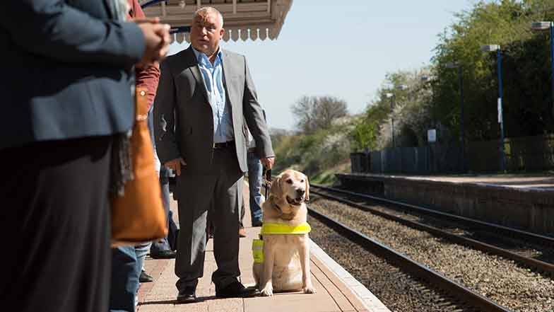 A guide dog owner with his dog, and other travellers, waiting on the platform for their train.