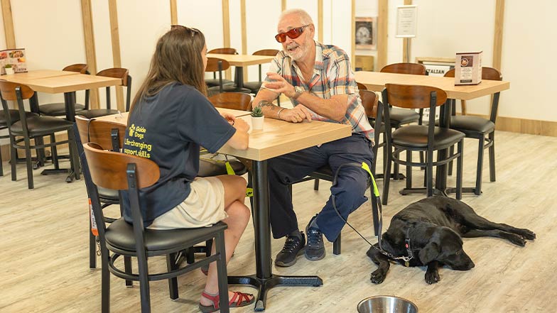 A guide dog owner chats to a Guide Dog Mobility Specialist, in a café, with his guide dog beside him.