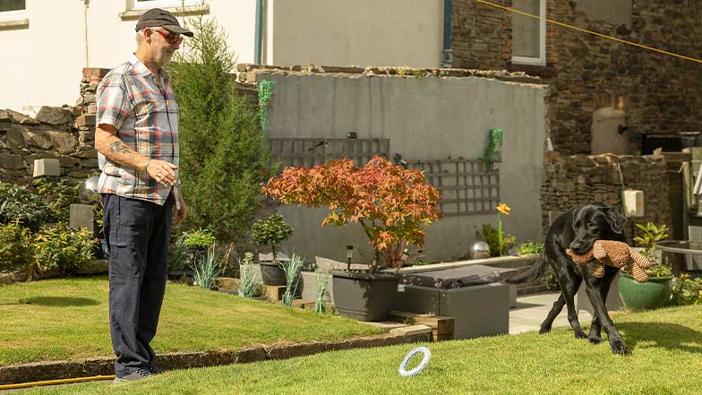 A guide dog owner and his guide dog play in their garden on a sunny day.