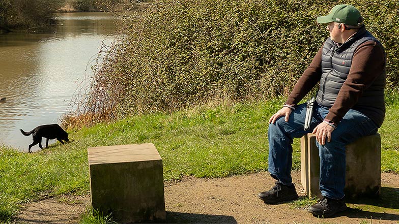 A long cane user sits on a seat in a sunny park. There is a lake in the background. To his right there is a black Labrador sniffing the grass.
