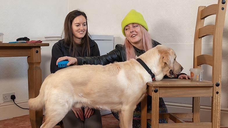 A guide dog performs a chin rest on a chair as her guide dog owner and a Guide Dog Mobility Specialist sit beside her.