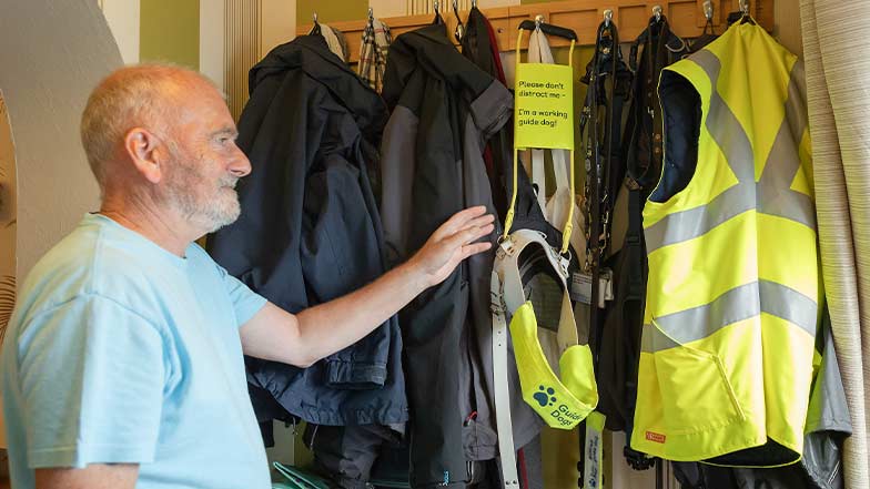 A guide dog owner reaches for a guiding harness hanging near his door, next to a reflective vest.