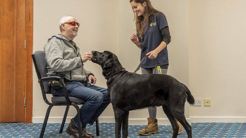A guide dog owners sits on a chair while giving his guide dog a food reward. A guide dog mobility specialist is stood nearby. They are all smiling.