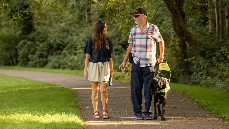 A Guide Dog Mobility Specialist walks alongside a guide dog owner and their guide dog in a sunny park.