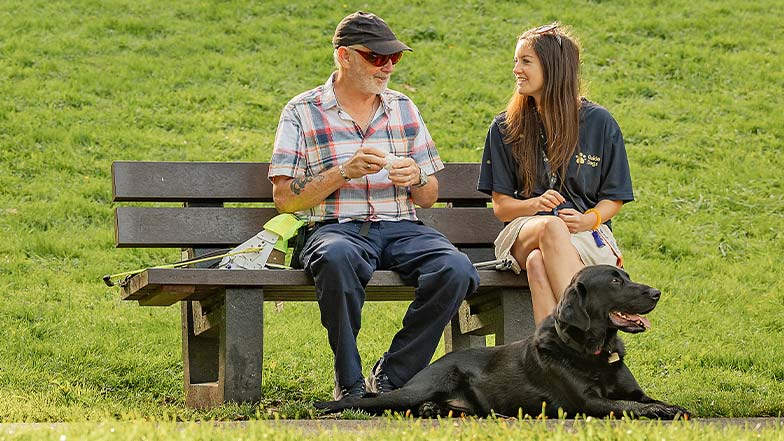 A guide dog owner sits on a park bench with his Guide Dog Mobility Specialist. His guide dog sits at his feet.