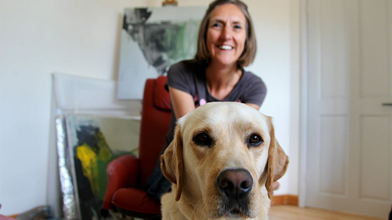 Artist Angela Charles sat on a chair with her guide dog Flynn in front of her looking to camera