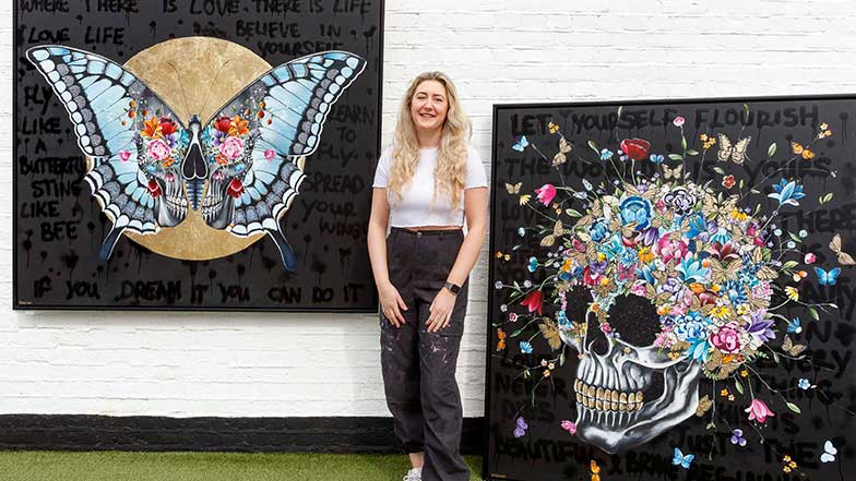 Artist Becky Smith standing beside two of her paintings, a butterfly and a skull