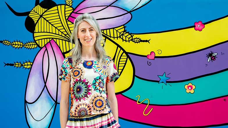 Artist Caroline Daly standing with a colourful bee painting behind her