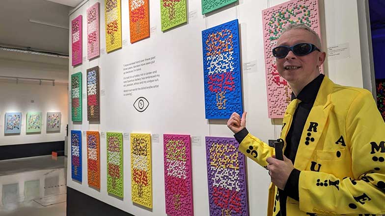 Artist Clarke Reynolds standing beside a wall showcasing several of his braille artworks