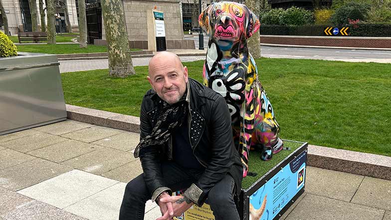 Artist Dom Pattinson sat on the plinth in front of his guide dog sculpture which is decorated in colourful street-style artwork with features such as butterflies, drips and lips.