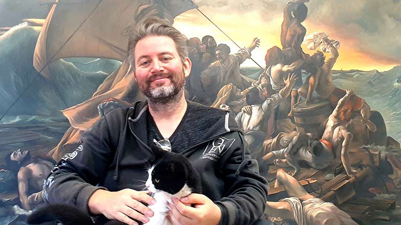 Artist Glen Brooks sat in front of a scenic wall painting with a black and white cat