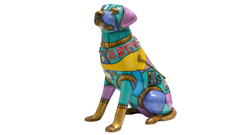 Painted guide dog sculpture with a multi-coloured metal futuristic patchwork robot dog design. 'Dog Brown' is written across the dog's shoulders with rivets detailing the words in braille..