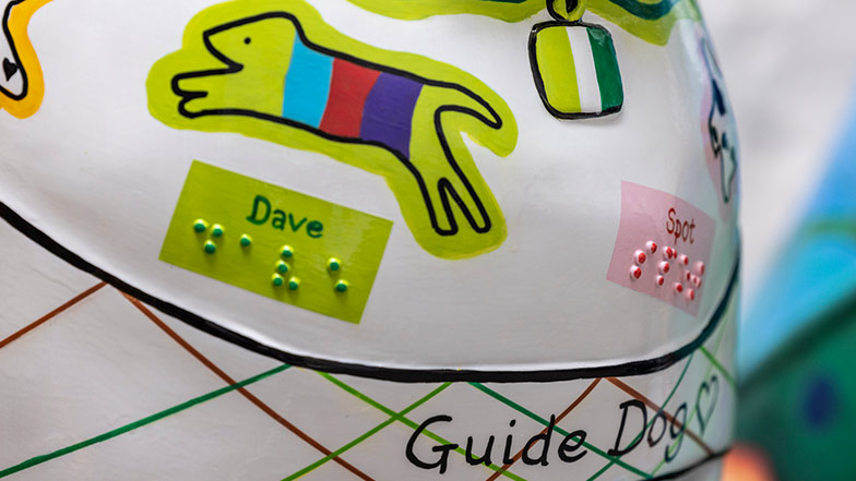 Close up of sculpture's chest showing a mini dog painted and its name 'Dave' written and in braille. On the front of the harness 'Guide Dog' is written.
