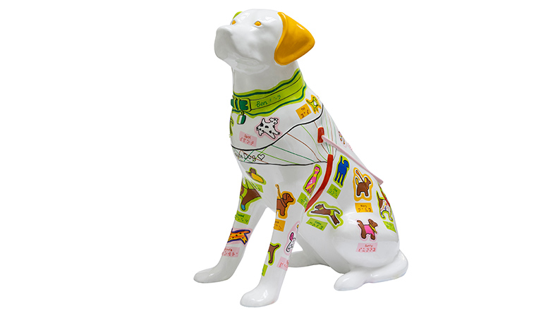 White guide dog sculpture with one green ear and one orange ear, plus brightly coloured mini dogs painted all over, featuring each dog's name in braille underneath.