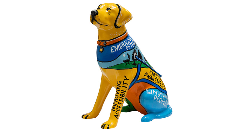Yellow guide dog sculpture with different phrases written on it. The dog has  blue, green and orange painted scenes of people walking with their guide dogs.