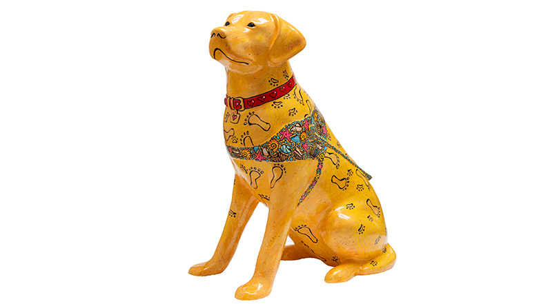 Yellow painted guide dog sculpture covered with sand and foot and paw prints spread out across the sand. The dog wears a shell-covered harness.