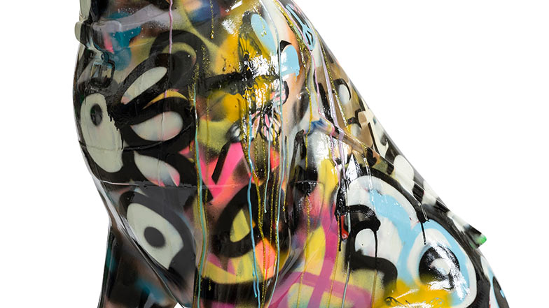 Close-up side view of the sculpture which is decorated with multi-coloured spray paint with motifs such as a butterfly and drips.