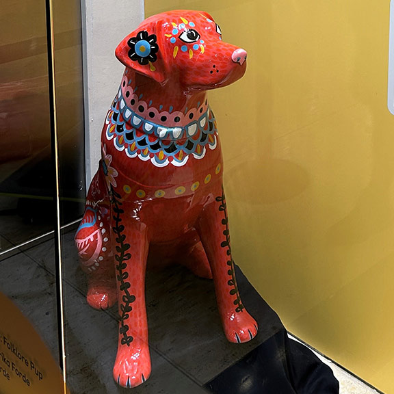Close up of mini guide dog sculpture painted red with folklore style detailing. Sculpture is sat in a window display in Canary Wharf.