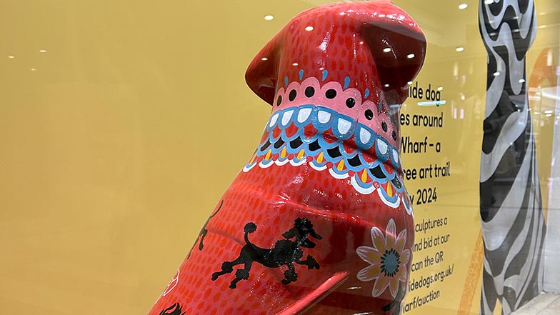 Back of the head and shoulders image of a mini guide dog sculpture painted red with a pink, blue, white and black folk style pattern around the neck and over the body.