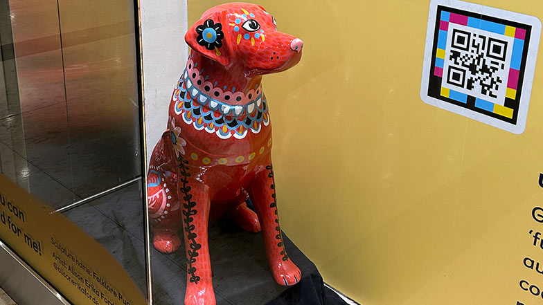 Front view of a guide dog sculpture sat in a window display. It is red and painted with black, blue, yellow and white folk style patterns.