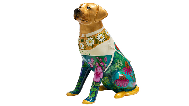 A yellow guide dog sculpture. The dog's chest is covered in white flowers. The dog's lower half is covered with vibrant flowers on a green background.