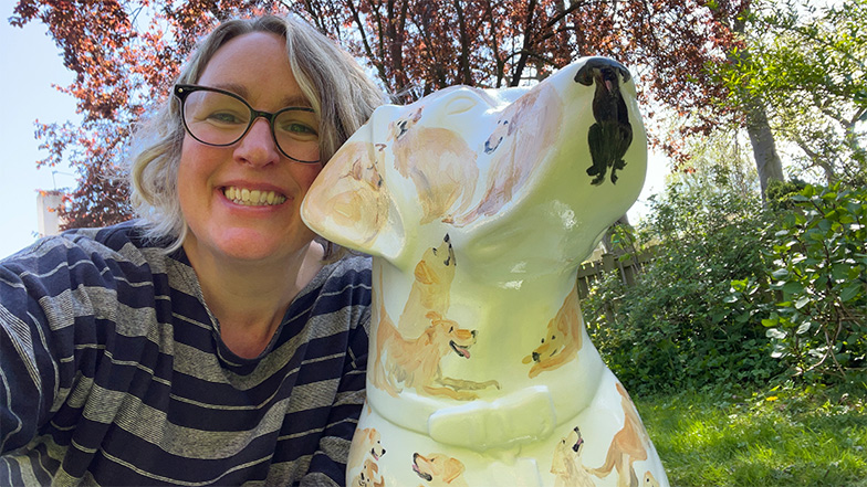 Head and shoulders image of artist Jenny Bloomfield next to her white guide dog sculpture painted with small yellow and black Labradors and golden retrievers.