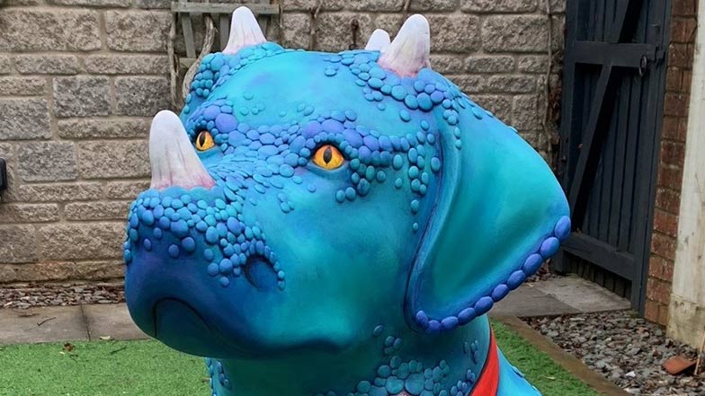 Close up of the sculpture's head which is painted blue and has tactile scales and horns growing out the dog's nose and head. It has yellow eyes.