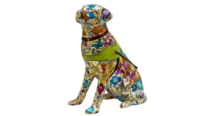 A gold guide dog sculpture with a multi-coloured floral and nature pattern including butterflies and birds. The dog has a red collar and a yellow harness with black handles.
