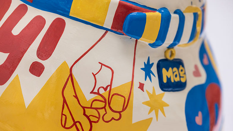 Close up of the dog's collar and chest. The collar is blue, white, yellow and red striped. There is a hand holding a treat painted on the chest. The collar tag reads 'Mag'.