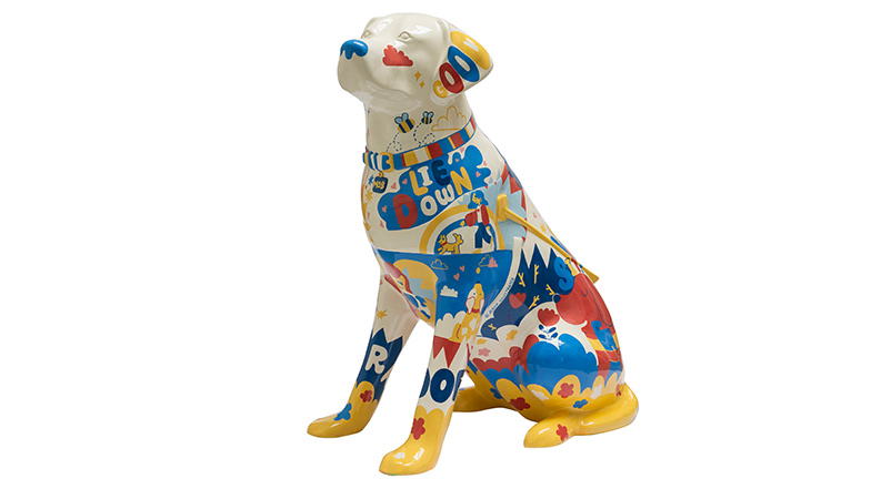 A cream coloured guide dog sculpture covered in a colourful pattern featuring dogs, their owners, toys and some dog-related words, such as 'lie down', 'woof' and 'good'.
