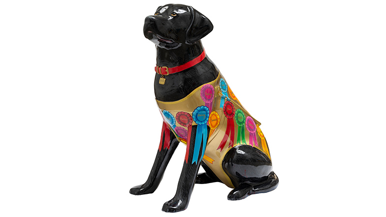 A black guide dog sculpture wearing a red collar and a gold dog coat with different coloured rosettes. The rosettes contain words that describe people's dogs, such as, loyal, trust and partner.