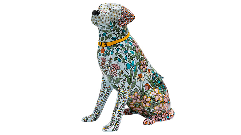 A guide dog sculpture painted light blue and is covered in flowers and plants. Hidden in the foliage are illustrations of dogs. 
